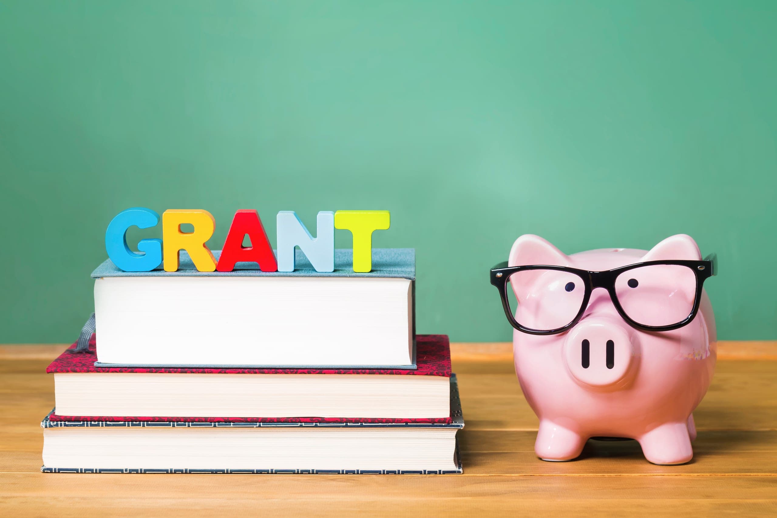 A wooden table displays a piggy bank and books, with a green board in the background, illustrating K-12 school safety grants.